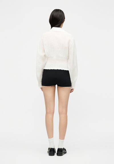 Femme portant un chemisier blanc à manches longues avec taille et poignets froncés, short noir aux bords effilochés, chaussettes blanches et mocassins noirs, vue de dos.