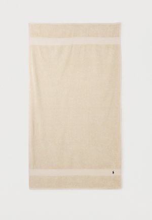Serviette rectangulaire beige avec deux bandes horizontales crème près des bords et un petit logo brodé bleu foncé dans le coin inférieur droit.