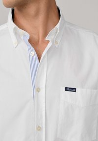 Chemise blanche à boutons avec un col intérieur rayé bleu et blanc, une poche avant avec une étiquette marine, et des boutons crème clair.