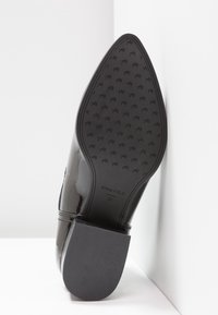 Scarpa alta in vernice nera con punta affusolata. La suola presenta una trazione strutturata con un motivo a logo e un tacco rettangolare.