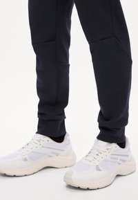Person iført hvide HUGO sneakers og sorte tætsiddende bukser står på en ensfarvet hvid baggrund.