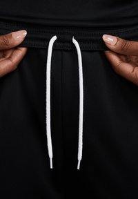 Pantaloni sportivi neri con una texture liscia, dotati di un cordino bianco e dettagli cuciti in vita. Mani tengono la vita.