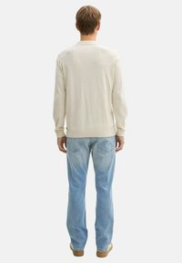 Beige pulóver bordázott textúrával és hosszú ujjal, világoskék farmerrel párosítva. A nadrág klasszikus öt zsebes dizájnnal rendelkezik.