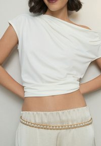 Valkoinen yhden olkapään toppi laskostetulla kankaalla, yhdistetty korkeavyötäröisiin, kermanvärisiin housuihin, joissa on kultainen helmikoristeinen ketjuvyö.