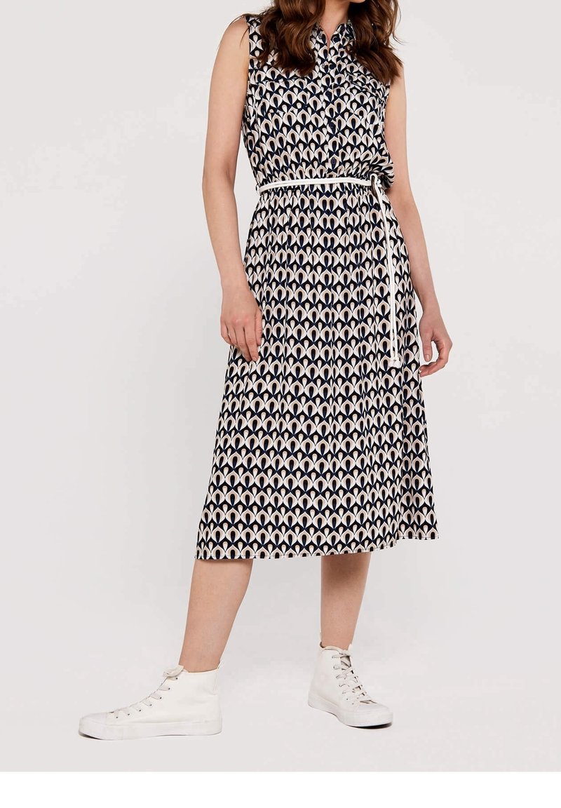 Femme portant une robe midi sans manches à motifs avec une ceinture blanche et des baskets montantes blanches, debout devant un fond uni.