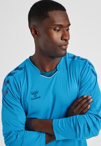 Lichtblauw sportshirt met lange mouwen en een ronde hals, met contrasterende marineblauwe strepen op de schouders en een klein logo op de borst.