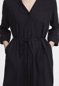 Schwarzes Hemdkleid mit V-Ausschnitt, langen Ärmeln und gebundenem Taillenbündchen. Hergestellt aus weichem Material und mit zwei Seitentaschen ausgestattet.