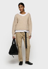 Beige gebreide trui met een V-hals, gelaagd over een witte shirt. Lichtbruine broek met een zwart banddetail. Zwarte tas en schoenen.