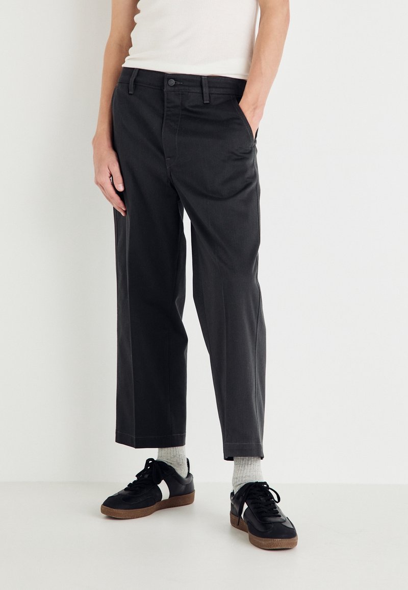 Personne portant un pantalon noir droit, des chaussettes blanches et des baskets noires avec des rayures blanches, debout avec une main dans la poche.