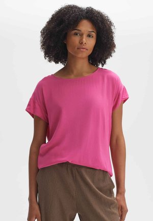 Blusa rosa a maniche corte realizzata in tessuto leggero, con scollatura a barca e vestibilità rilassata. Indossata con pantaloni marroni a trama.