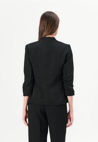 Černý blazer s přiléhavým střihem, dlouhými rukávy vyhrnutými nahoru a vysokým límcem. Hladká textura a minimalistický design bez viditelných zapínání.