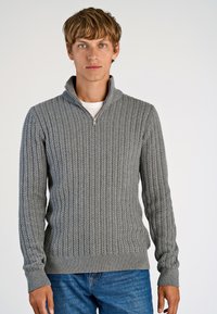 Szary sweter dzianinowy z wysokim kołnierzem, posiadający zamek błyskawiczny, ribbową teksturę i długie rękawy. Noszony z niebieskimi dżinsami.