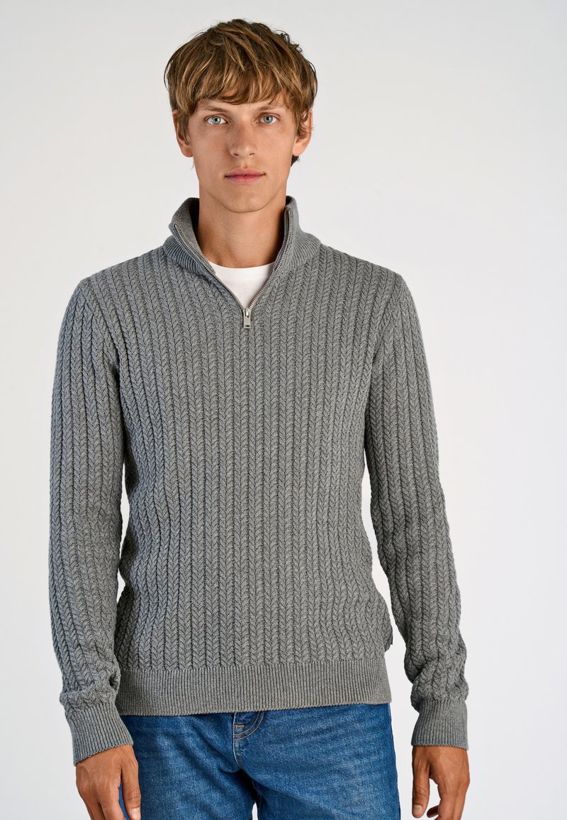 Szary sweter dzianinowy z wysokim kołnierzem, posiadający zamek błyskawiczny, ribbową teksturę i długie rękawy. Noszony z niebieskimi dżinsami.