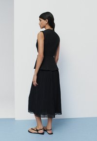 Svart peplumtop paired med en lång, plisserad svart kjol. Outfiten har ett texturerat tyg och subtila lager, tillsammans med platta sandaler.