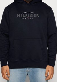 Felpa blu navy con tasca a marsupio frontale, cappuccio con coulisse e logo bianco "HILFIGER" che include l'anno di fondazione e un accento rosso.