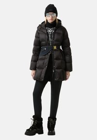 Černá péřová bunda se staženým pasem, v kombinaci s vzorovaným svetrem, černými legínami a koženými botami s kožešinovou podšívkou. Doplněno čepicí a slunečními brýlemi.