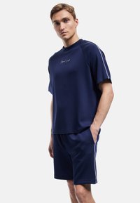 Sættet i marineblå bomuld til atletisk brug består af en t-shirt med korte ærmer og rund hals samt shorts med sidelommer og hvide sidelister.