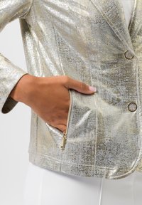 Metallic gouden en zilveren blazer met een gestructureerde afwerking, voorzien van een ritszak en ronde zilveren knopen. Op maat gemaakte design met notch-ails.