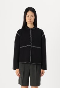 Zwarte gebreide jas met contrasterende witte stiksels. Bevat een ronde hals, lange mouwen en knopen aan de voorkant. Draagt over grijze shorts.