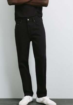 Homme portant un jean noir et des baskets blanches, debout les bras croisés devant un fond clair uni.