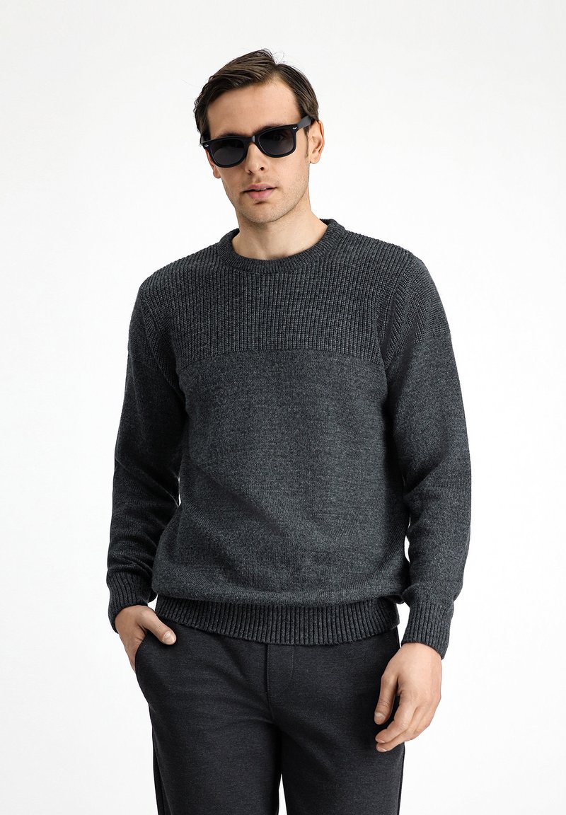 Jersey de punto gris oscuro con textura acanalada, cuello redondo y mangas largas, combinado con pantalones gris oscuro. El actor lleva gafas de sol negras.