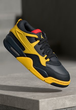 Czarno-żółty sportowy but na sznurówki z czarnymi sznurowadłami i czerwonym językiem wewnętrznym, zawieszony nad szarą kamienną powierzchnią.