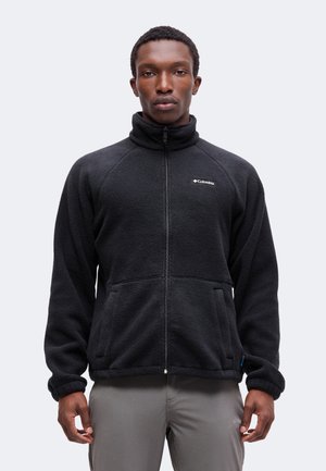 Homme adulte portant une veste polaire noire zippée intégralement avec un col montant et un pantalon gris, debout devant un fond clair uni.