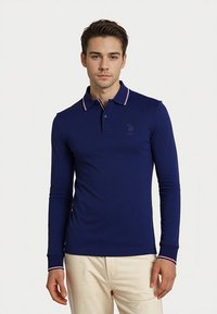 Mladý muž oblečený v úzkom navy modrom polokošeli s dlhým rukávom, s červeným a bielym lemovaním, stojaci na neutrálne svetlom pozadí.