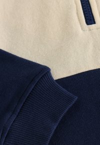 Zwei Stoffabschnitte: einer beige mit einem Reißverschluss, der andere dunkelmarine. Beide haben eine gerippte Textur. Einfaches Farbblocking-Design.