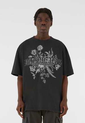 Sort oversized t-shirt i bomuld, med et hvidt blomster grafik og tekstdesign på fronten. Korte ærmer, rund hals.