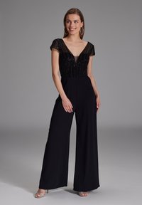 Tuta nera con scollatura a V profonda e maniche corte trasparenti decorate con perline. Il tessuto è liscio, fluisce con gambe larghe e un corpetto aderente.