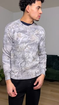 Szürke hosszú ujjú pulóver texturált anyagból, levelekkel díszítve. Kerek nyakkal és testre szabott ujjal rendelkezik, fekete nadrággal párosítva.