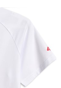Section de chemise blanche à manches courtes avec couture visible et petit logo triangulaire rouge près de l'ourlet de la manche.