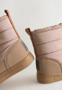 Quilted beige Knöchelstiefel mit tanfarbenen Gummisohlen, genähten Akzenten und Zuglaschen auf der Rückseite; glatte Stofftextur mit subtiler glänzender Oberfläche.