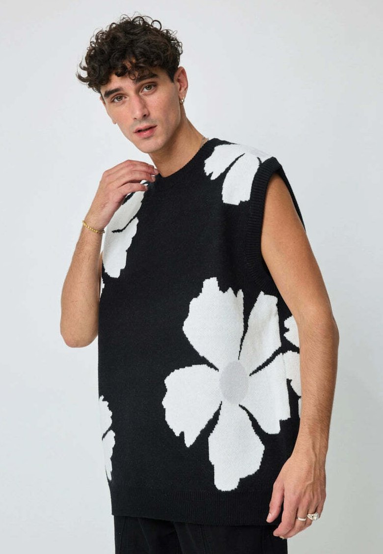 Homme avec des cheveux bouclés portant un gilet sans manches noir avec de grands motifs de fleurs blanches, touchant le col sur fond gris uni.