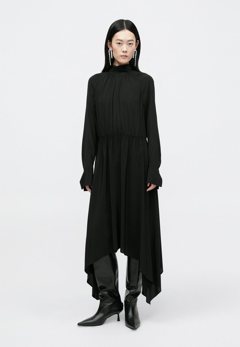 Vestido negro de manga larga con cintura fruncida, escote alto y dobladillo asimétrico; combinado con botas ajustadas hasta la rodilla en negro.