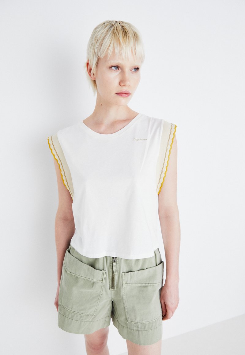 Hvit bomull t-skjorte med bølgede ermekanter i gul og beige, sammen med grønne shorts med glidelås og store lommer.