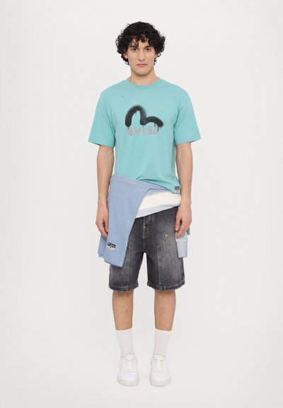 T-shirt turchese con stampa grafica, maglione azzurro chiaro annodato in vita, pantaloni shorts di jeans scuri, calze bianche, sneakers bianche.