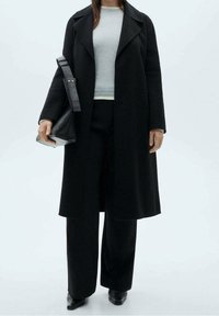 Trench-coat en laine noire avec de larges revers, associé à un pull gris clair et un pantalon noir à jambes larges. Tient un petit sac noir.