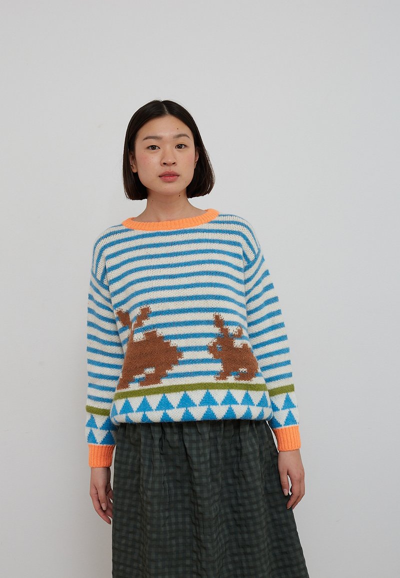 Femme portant un pull rayé bleu et blanc avec des motifs de lapins marron et des accents orange, associé à une jupe à carreaux vert foncé.
