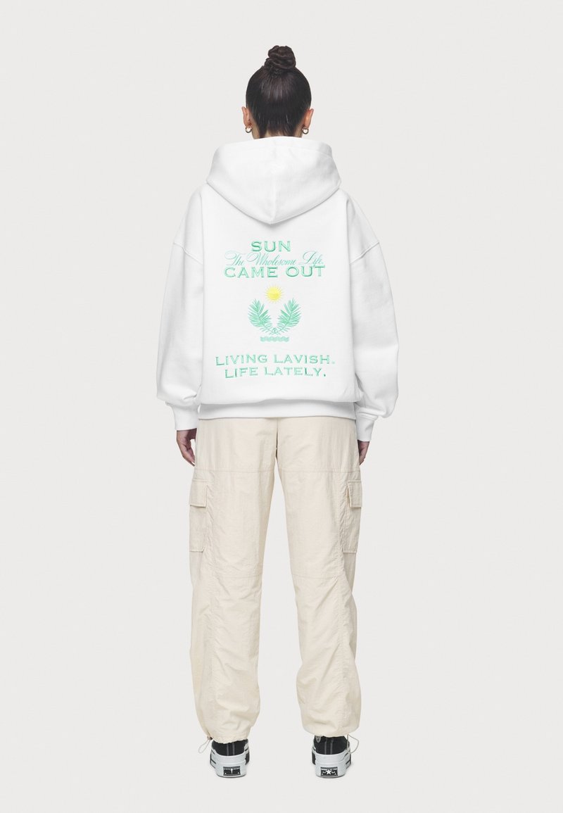 Personne portant un sweat à capuche blanc avec un texte vert et un graphique de soleil dans le dos, pantalon cargo beige, baskets noires et blanches, debout, dos tourné.