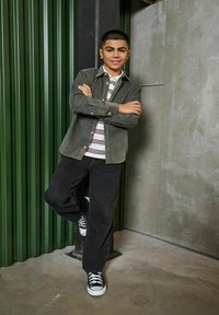 Groene corduroy button-up over een wit en gestreept T-shirt, gecombineerd met zwarte wijde jeans en zwarte sneakers, poserend tegen een groene achtergrond.