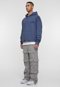 Blue cotton hoodie with a front pocket, layered over a white shirt. Gray cargo pants feature multiple pockets. White and gray sneakers.