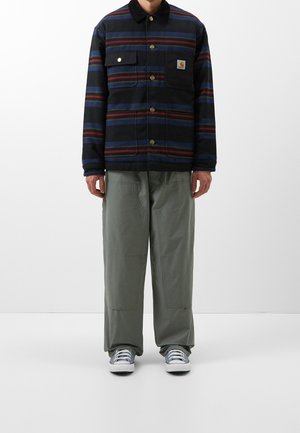 Uomo con giacca a righe nere, blu e rosse con bottoni, pantaloni grigi larghi e sneakers grigie, in piedi davanti a uno sfondo bianco uniforme.