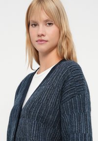 Tânără cu păr blond, îmbrăcată într-o jachetă tricotată texturată de culoare albastră închisă, purtând un top alb, așezată cu fața ușor spre lateral, pe un fundal simplu.