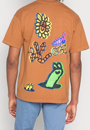 Person iført en brun T-shirt med farverige, abstrakte insekts- og blomsterdesigns på ryggen, kombineret med lyseblå bukser.