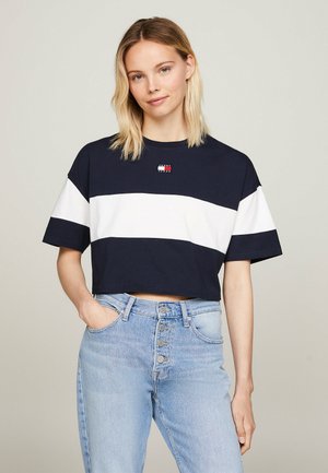 Mladá žena s blond vlasmi, ktorá má na sebe námornícky modré a biele pruhované krátke tričko a svetlomodré rifle s vysokým pásom, stojí s jednou rukou v vrecku.