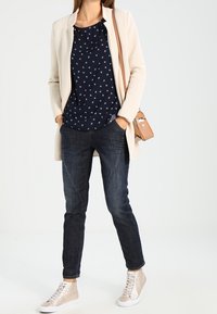 Beige lång kofta över en marinblå blus med vita mönster, ihop med mörka jeans och beige högskaftade sneakers. Liten tan färgad axelväska.