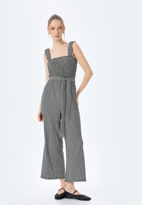 Grå och vit rutig jumpsuit med smockad byst, vida ben och bälte; har tjocka axelremmar och texturerad tyg.