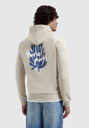 Man met krullend haar, gekleed in een beige hoodie met een blauw-koraal ontwerp en de tekst "Pure Earth" op de achterkant, gecombineerd met lichtblauwe spijkerbroek.